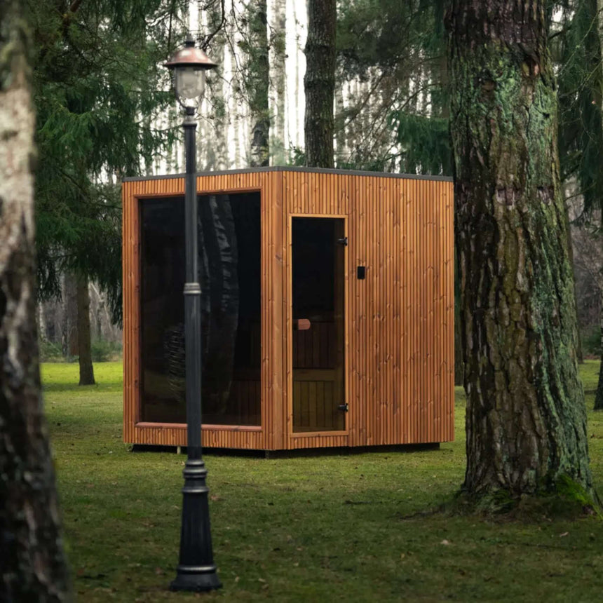 Celsium Jonas Classic outdoor sauna with vertical ThermoWood cladding and a glass door, seen past a lamp post between tree trunks