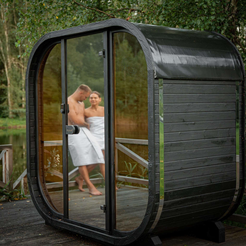 Dark Celsium Ula Mini outdoor sauna with a full glass front on a lakeside deck, with a couple in towels reflected in the glass