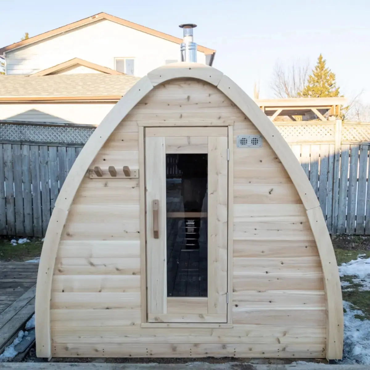 Front exterior view of a Dundalk LeisureCraft Canadian Timber MiniPOD Sauna (CTC77ME) in a snowy backyard, showing the main door and wood stove chimney.