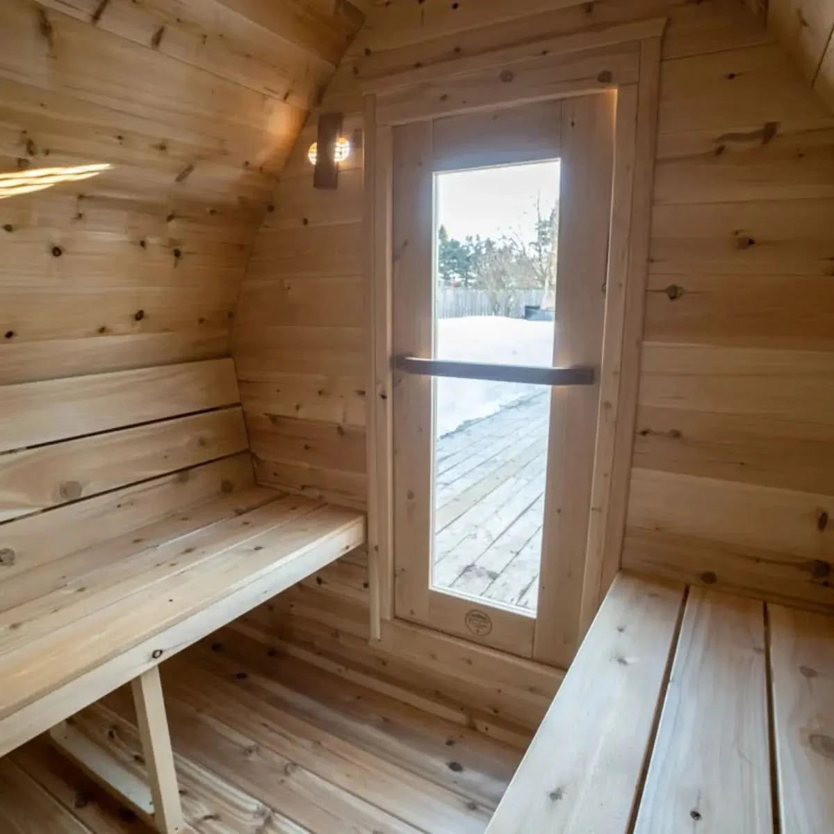 An interior view of a Dundalk LeisureCraft CT MiniPOD Sauna (CTC77ME), looking out through the door with a wooden handle towards a snowy backyard.