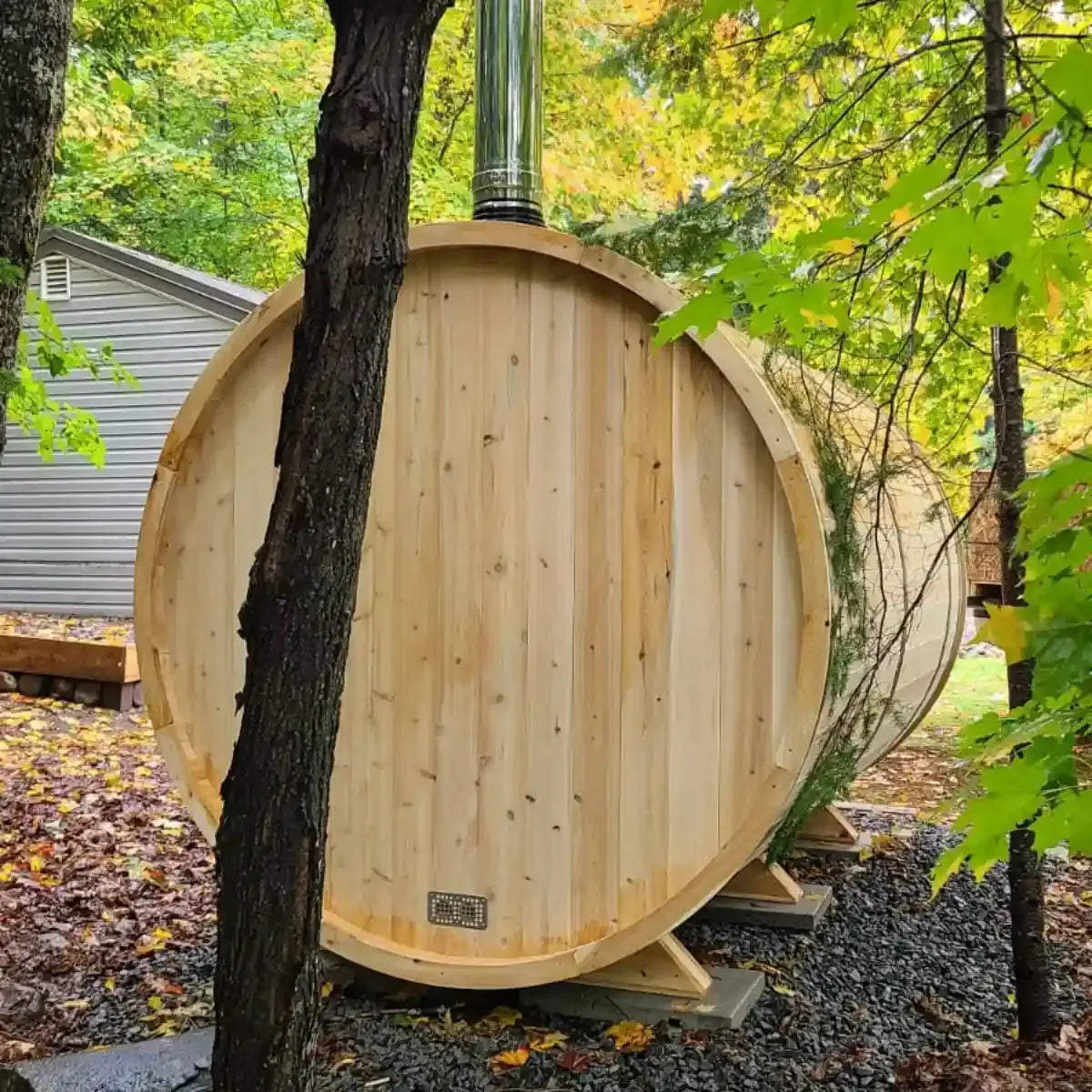 An exterior view of the Dundalk Leisure Craft CT Tranquility MP Barrel Sauna (CTC2345MPE) with an installed wood-burning stove and chimney pipe, surrounded by trees.
