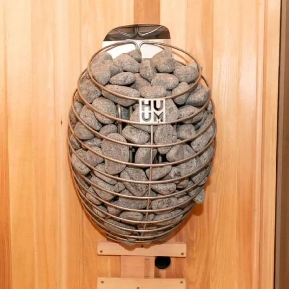 A close-up of the HUUM DROP electric sauna heater in its droplet-inspired metal frame, filled with round sauna stones and mounted on a wood-paneled wall.