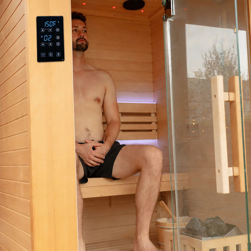 Man seated on the upper bench of the Revive 2 Person Traditional Sauna, with ceiling lights and the heater beside him