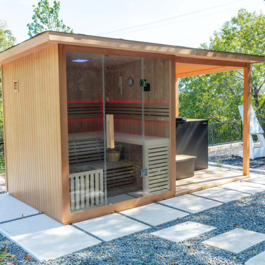 Revive 4 Person Traditional Sauna, sold separately, with red LED lighting inside and the deck extension on its right, outdoors on concrete pavers, with the Revive Luxury Plunge and its step, also sold separately, under the covered deck