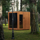 Celsium Jonas Classic outdoor sauna with vertical ThermoWood cladding and a glass door, seen past a lamp post between tree trunks