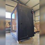 Side wall of the Celsium Jonas Mini outdoor sauna with a flush wooden door, dark ThermoWood cladding and lighting along the roofline