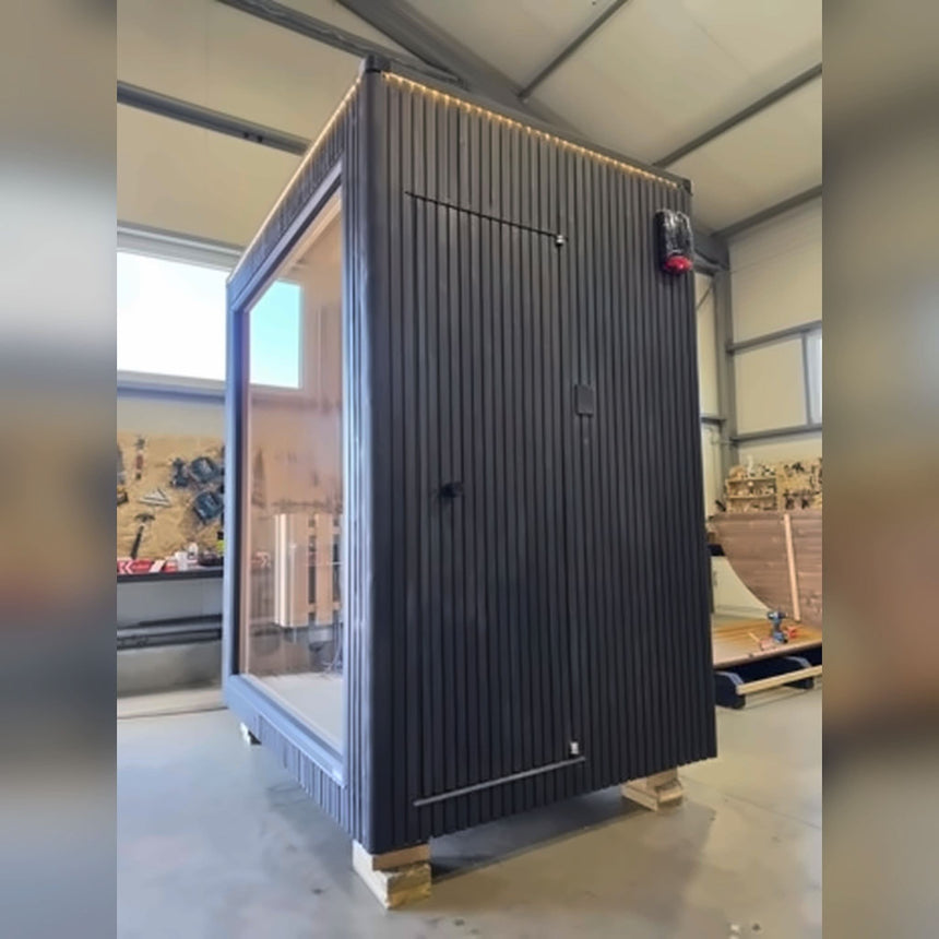 Side wall of the Celsium Jonas Mini outdoor sauna with a flush wooden door, dark ThermoWood cladding and lighting along the roofline