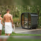 Couple in towels walking toward a dark Celsium Ula Mini outdoor sauna on a lakeside deck