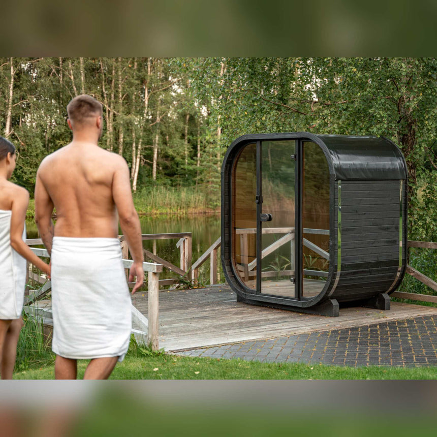 Couple in towels walking toward a dark Celsium Ula Mini outdoor sauna on a lakeside deck