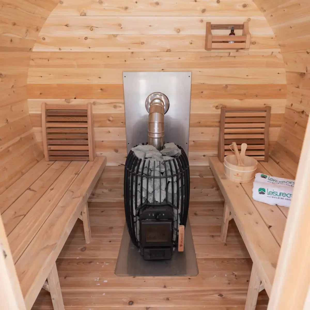 The interior of a Dundalk LeisureCraft CT Elation Sauna (CTC2424E), showing the wood-burning heater, two long benches, and a bucket with ladle.