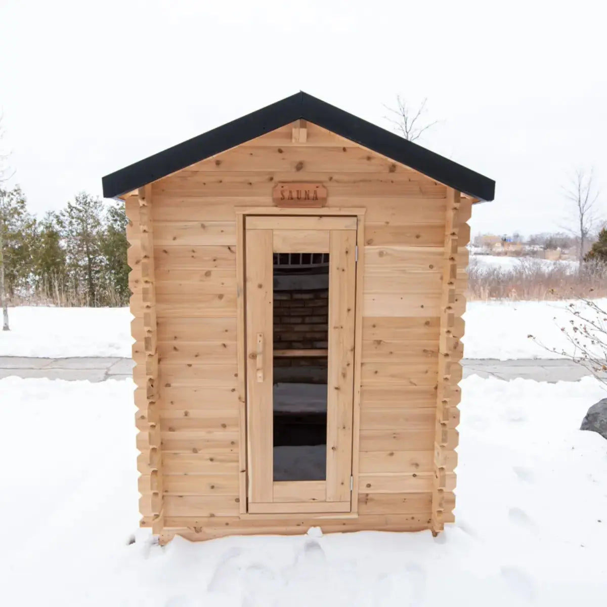Front exterior of a Dundalk LeisureCraft Canadian Timber Granby Cabin Sauna (CTC66E) in a snowy winter setting, showing the glass door.