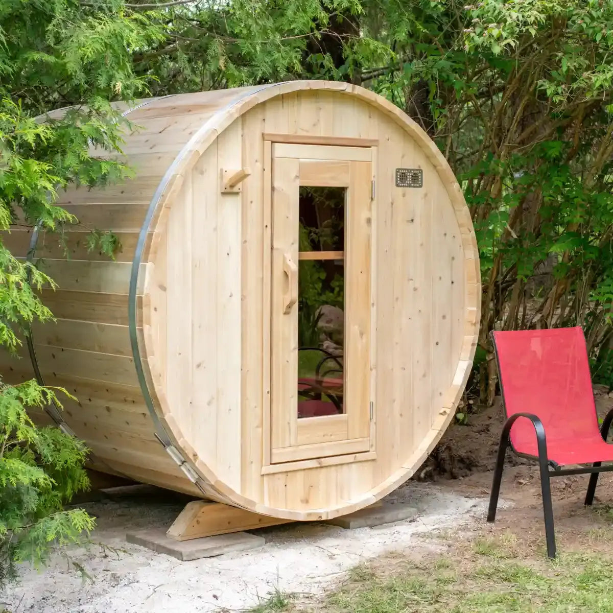 A full exterior view of a Dundalk LeisureCraft Canadian Timber Harmony Barrel Sauna (CTC22E) with a red chair next to it, showing the curved design and entrance.