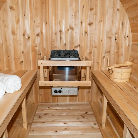 A detailed interior view of the Dundalk Leisure Craft CT Serenity Barrel Sauna (CTC2245E), highlighting the electric sauna heater, hot stones, a wooden bucket, and a ladle.