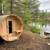 A beautiful lifestyle shot of the Dundalk Leisure Craft CT Serenity Barrel Sauna (CTC2245E) with an optional shingle roof, positioned on a gravel pad near a tranquil lake and wooded area.