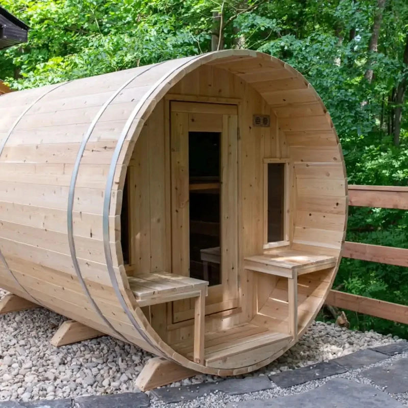 A beautiful exterior lifestyle shot of the Dundalk Leisure Craft CT Tranquility Barrel Sauna (CTC2345W) with an outdoor porch and side benches, set up in a backyard surrounded by trees.