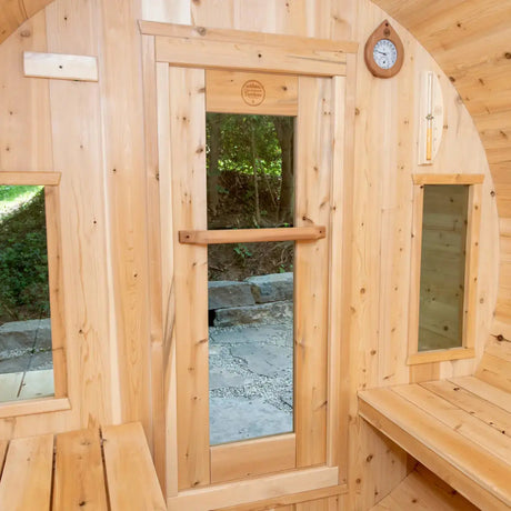 An interior view of the Dundalk Leisure Craft CT Tranquility Barrel Sauna (CTC2345W), showing the benches, a round window, and a vertical glass door with the brand logo.