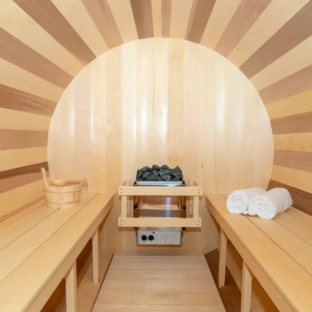An interior view of the Dundalk Leisure Craft CT Tranquility MP Barrel Sauna (CTC2345MPE), showing the sauna heater with stones, cedar benches, and a wooden bucket and ladle.