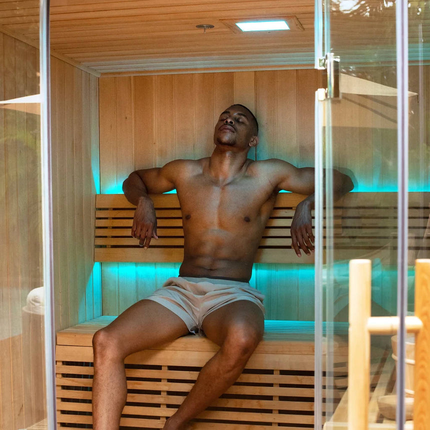 Man seated on the bench of the Revive 4 Person Traditional Sauna with teal LED lighting above and below the backrest