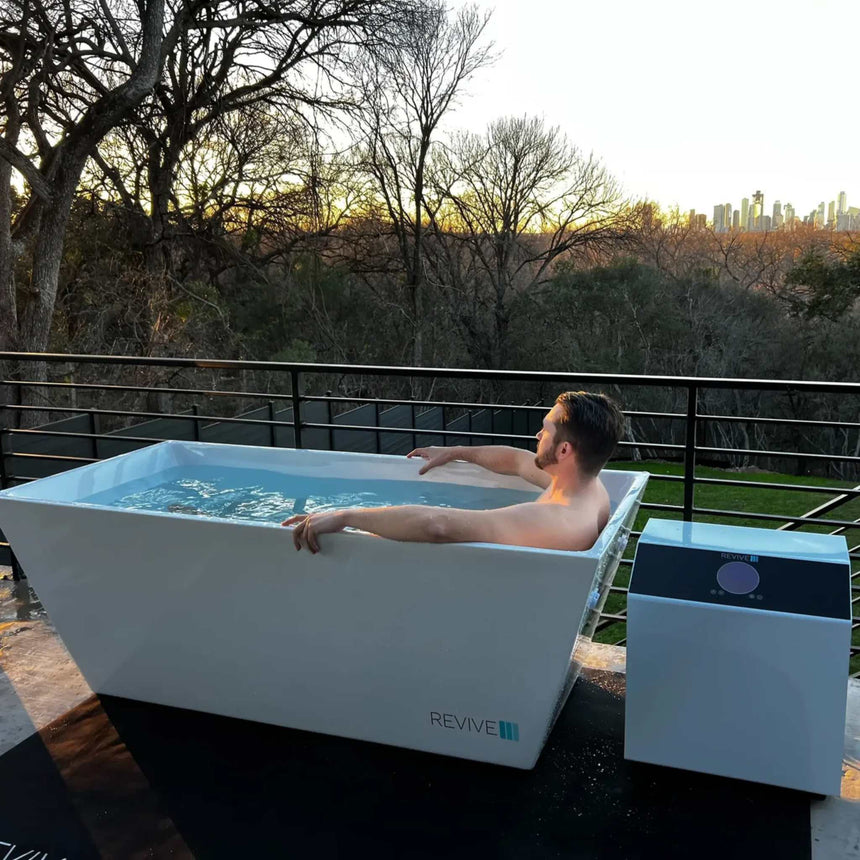 Man soaking in the Revive Acrylic Plunge on a terrace at sunset, with the chiller beside the tub, on the optional protective mat