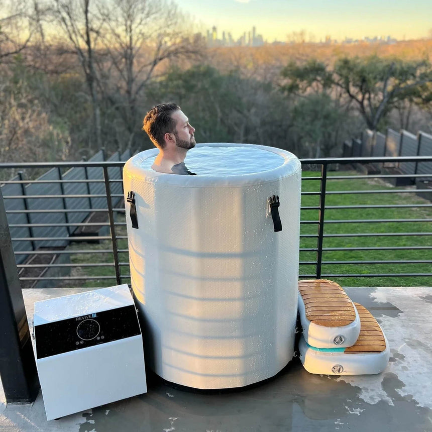 Man soaking in the Revive Inflatable Barrel Plunge on a terrace at sunset, with the chiller and step beside it