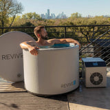 Man soaking in the Revive Inflatable Pod Plunge on a terrace overlooking a city skyline, with the chiller beside the tub