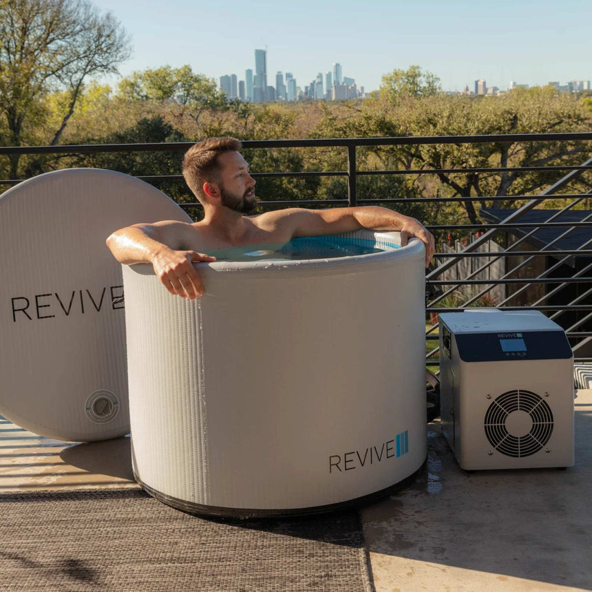 Man soaking in the Revive Inflatable Pod Plunge on a terrace overlooking a city skyline, with the chiller beside the tub