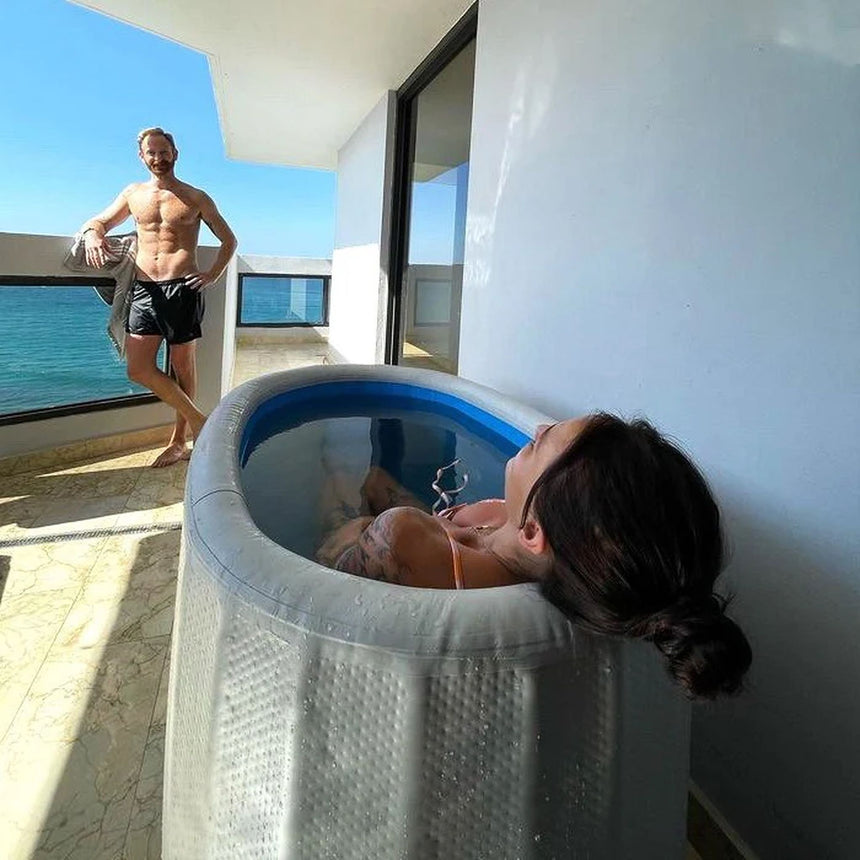 Woman soaking in the Revive Inflatable Tub on a sunny balcony overlooking the ocean, with a man standing nearby